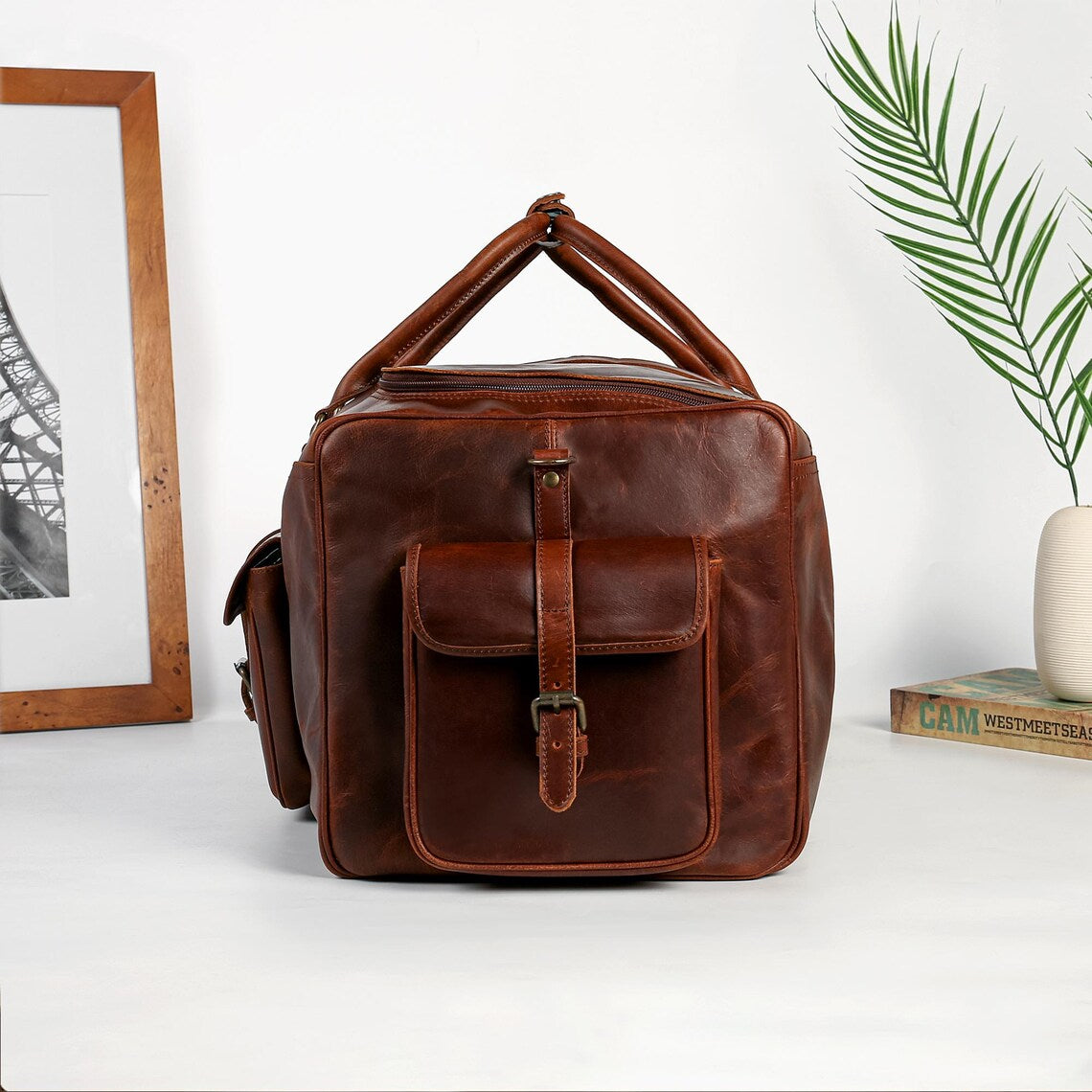 Brown leather bag on a white surface with a plant and framed picture in the background